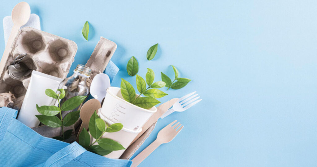 A blue background features a tote bag filled with eco-friendly items: wooden utensils, paper cups, a glass bottle, and green leaves.